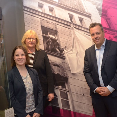 Zwei Frauen und ein Mann stehen vor einem Schwarz-Weiß-Foto. Es zeigt zwei Frauen, die eine weiße Fahne schwenken.