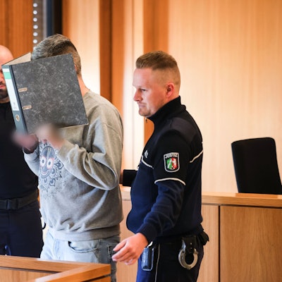 Der angeklagte Krankenpfleger hält sich einen Aktenordner vor sein Gesicht, während er von Justizangestellten zu seinem Platz gebracht wird.