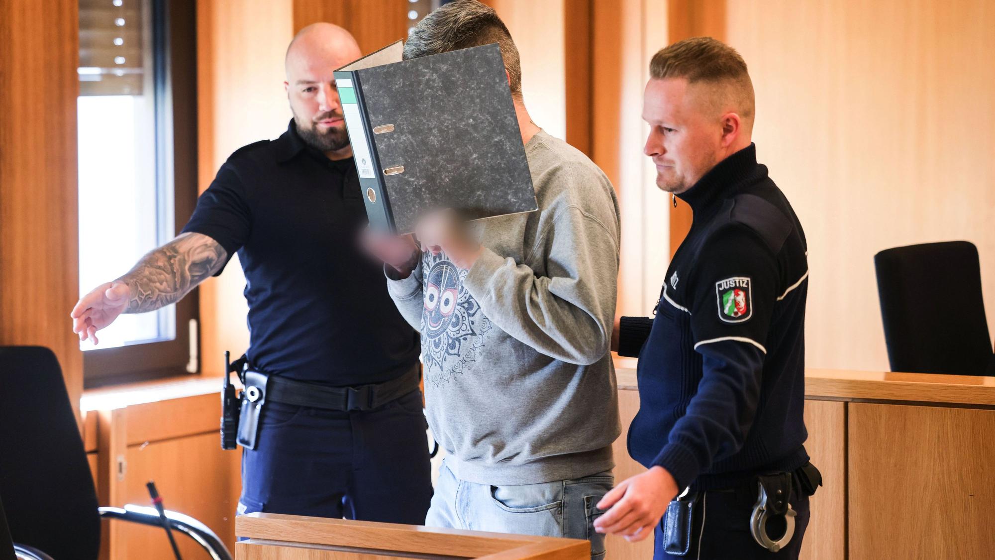 Der angeklagte Krankenpfleger hält sich einen Aktenordner vor sein Gesicht, während er von Justizangestellten zu seinem Platz gebracht wird.