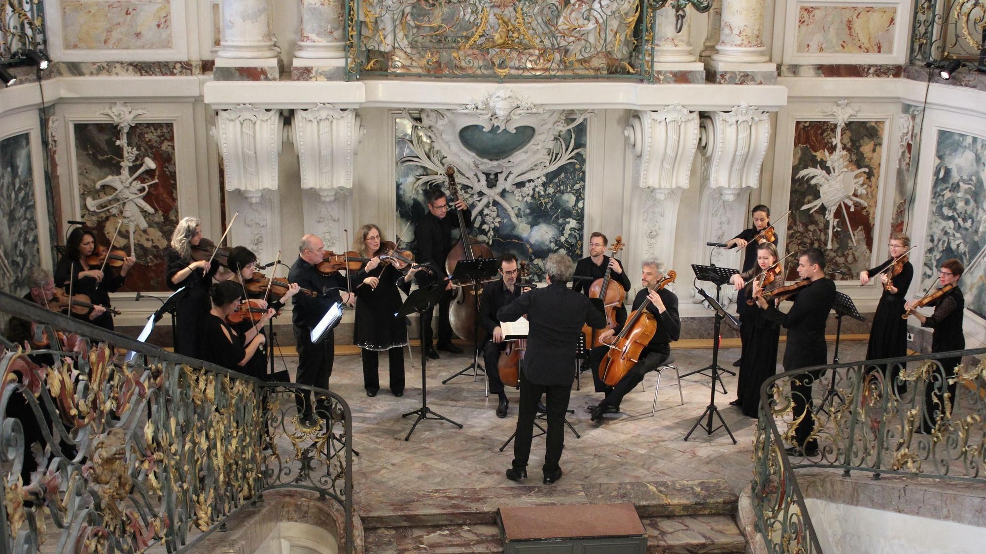 Zu sehen sind Musiker mit unterschiedlichen Instrumenten in einem mit Marmor verkleideten Treppenhaus eines Schlosses.
