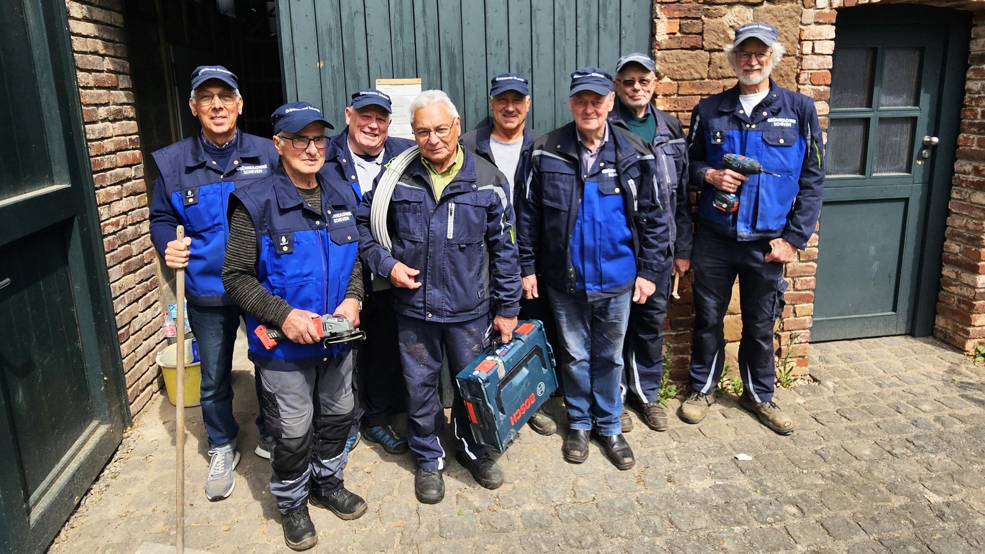 Eine Gruppe älterer Männer in blauer Arbeitskleidung. Einige halten Werkzeuge in den Händen.