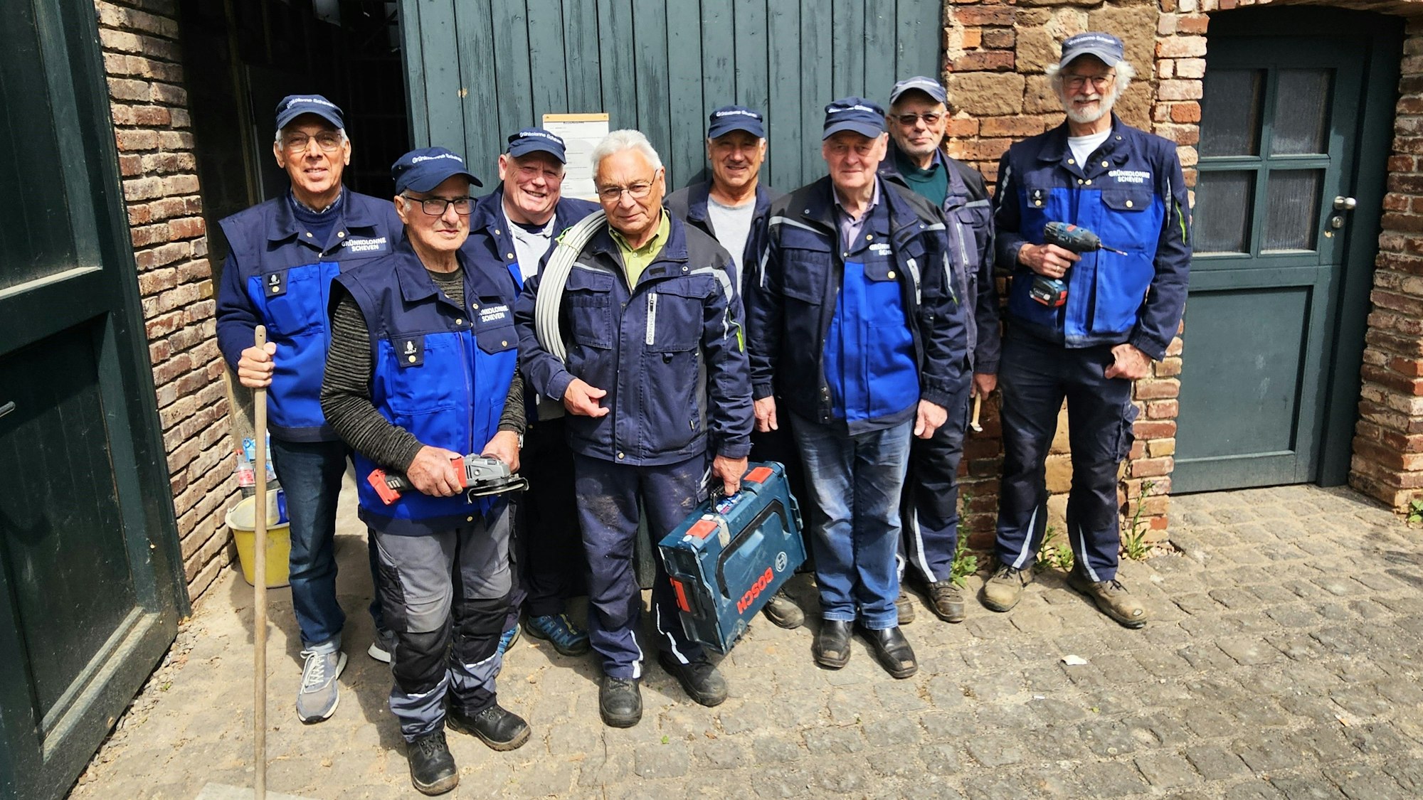 Eine Gruppe älterer Männer in blauer Arbeitskleidung. Einige halten Werkzeuge in den Händen.