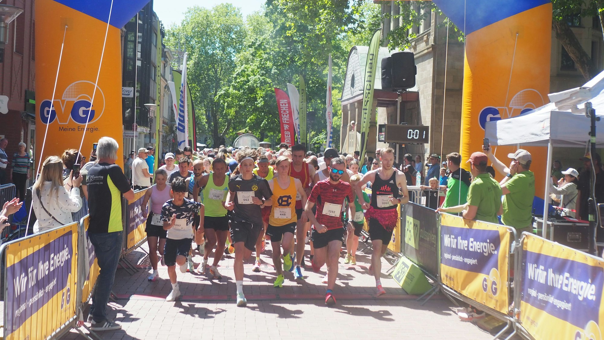 Die Läufer tragen bunte Trikots und Sportshirts und laufen gemeinsam los, links und rechts neben ihnen begrenzen Banden den Weg.