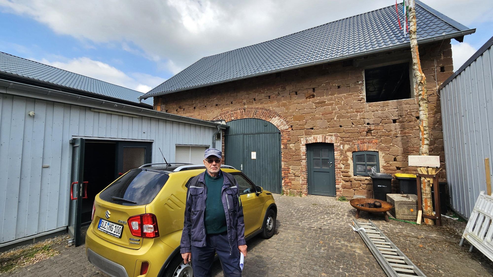 Hans Reiff mit Kappe und Arbeitskleidung steht neben einem Kleinwagen auf dem Hof vor einem aus Sandstein errichteten Scheunengebäude mit grünem Tor.