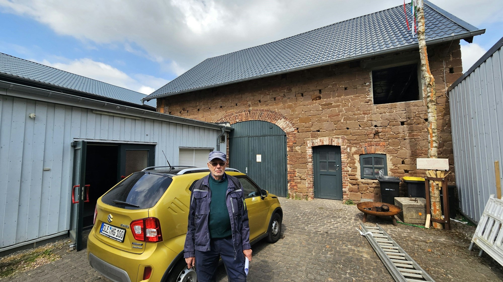 Hans Reiff mit Kappe und Arbeitskleidung steht neben einem Kleinwagen auf dem Hof vor einem aus Sandstein errichteten Scheunengebäude mit grünem Tor.
