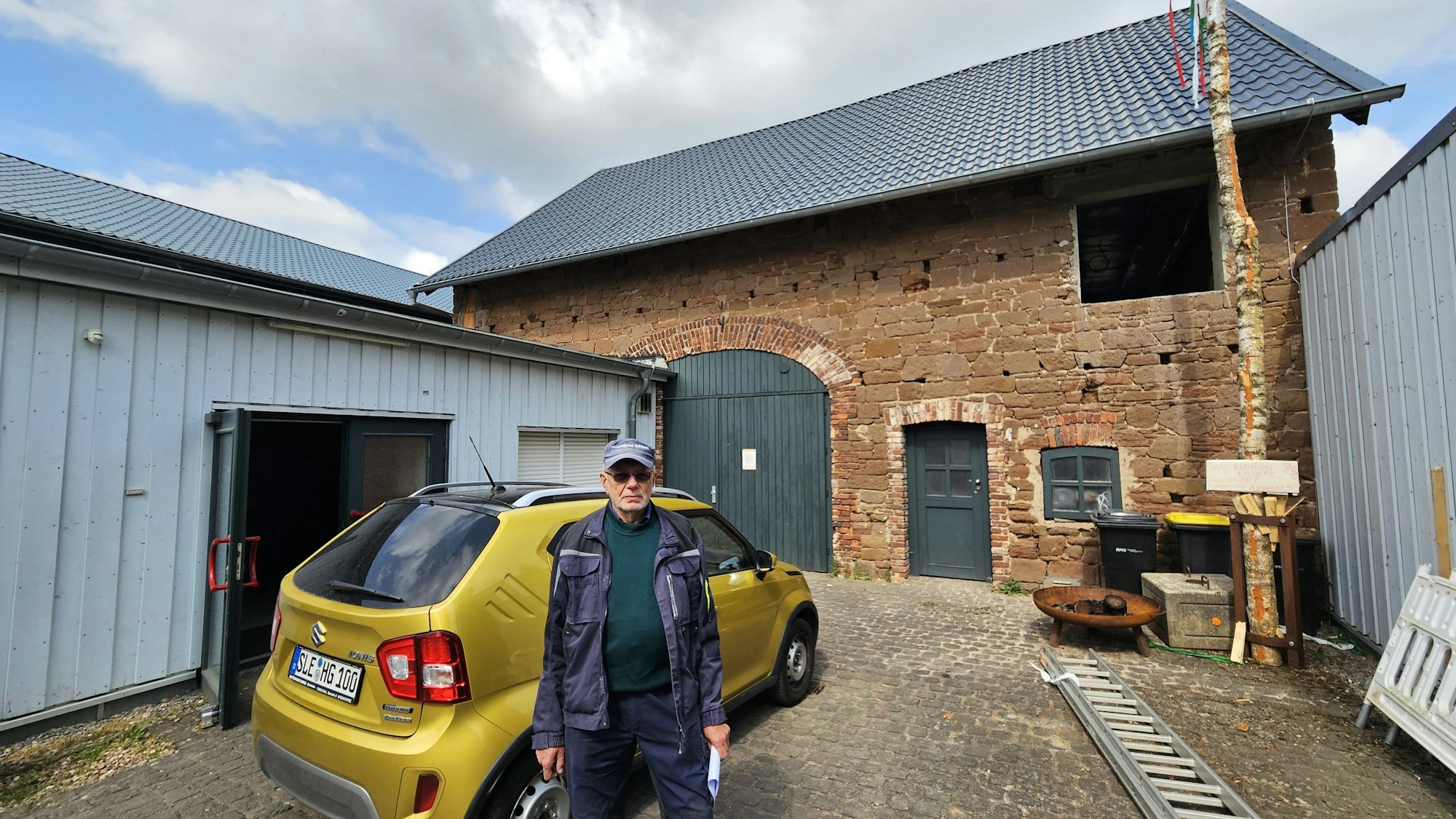 Hans Reiff mit Kappe und Arbeitskleidung steht neben einem Kleinwagen auf dem Hof vor einem aus Sandstein errichteten Scheunengebäude mit grünem Tor.