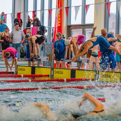 Zu sehen ist ein Schwimmbecken, im Vordergrund ein Schwimmer, im Hintergrund springt einer in das Wasser. Zahlreiche Zuschauer und weitere Schwimmer am Beckenrand sind ebenfalls zu sehen.