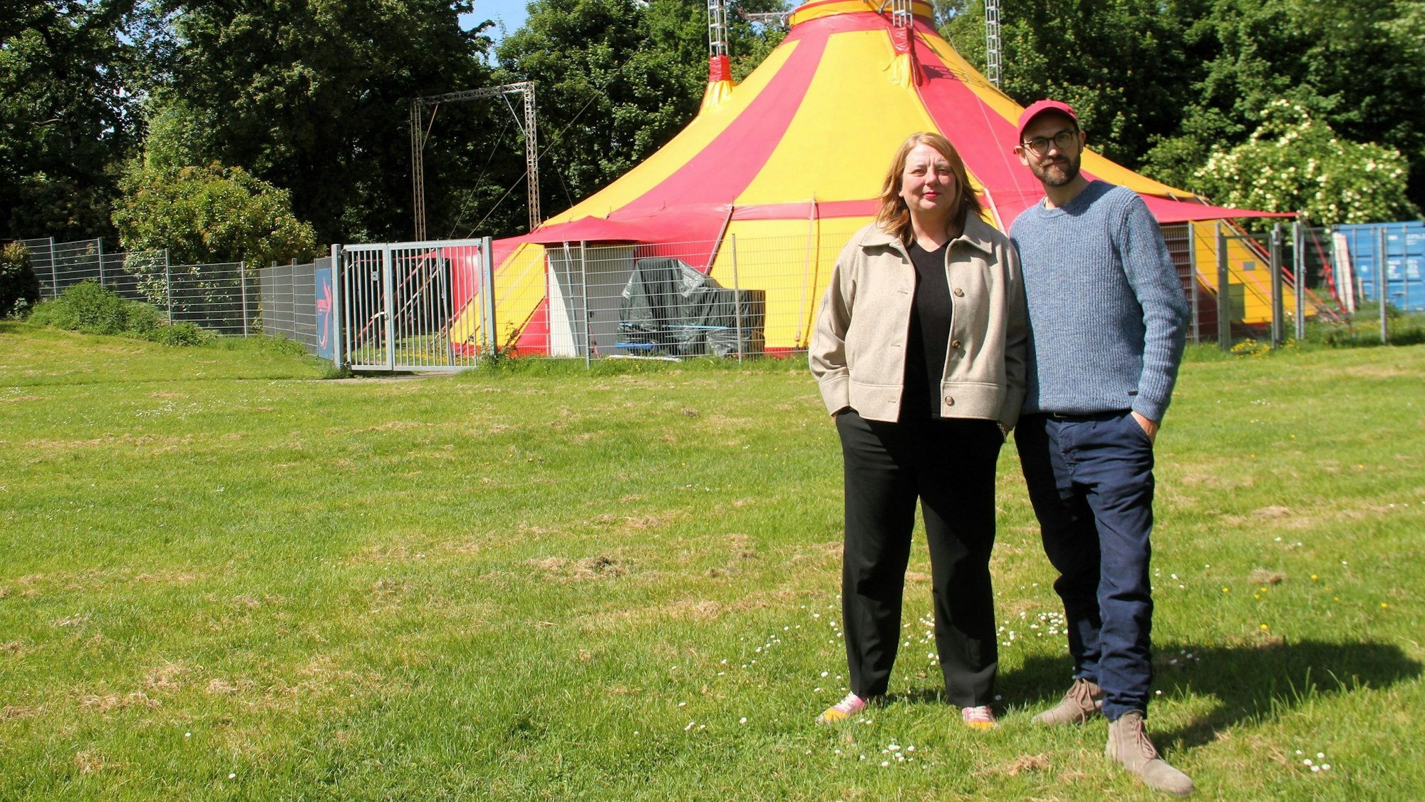 Eine Frau und ein Mann stehen nebeneinander auf einer Wiese vor einem gelb-roten Zirkuszelt.