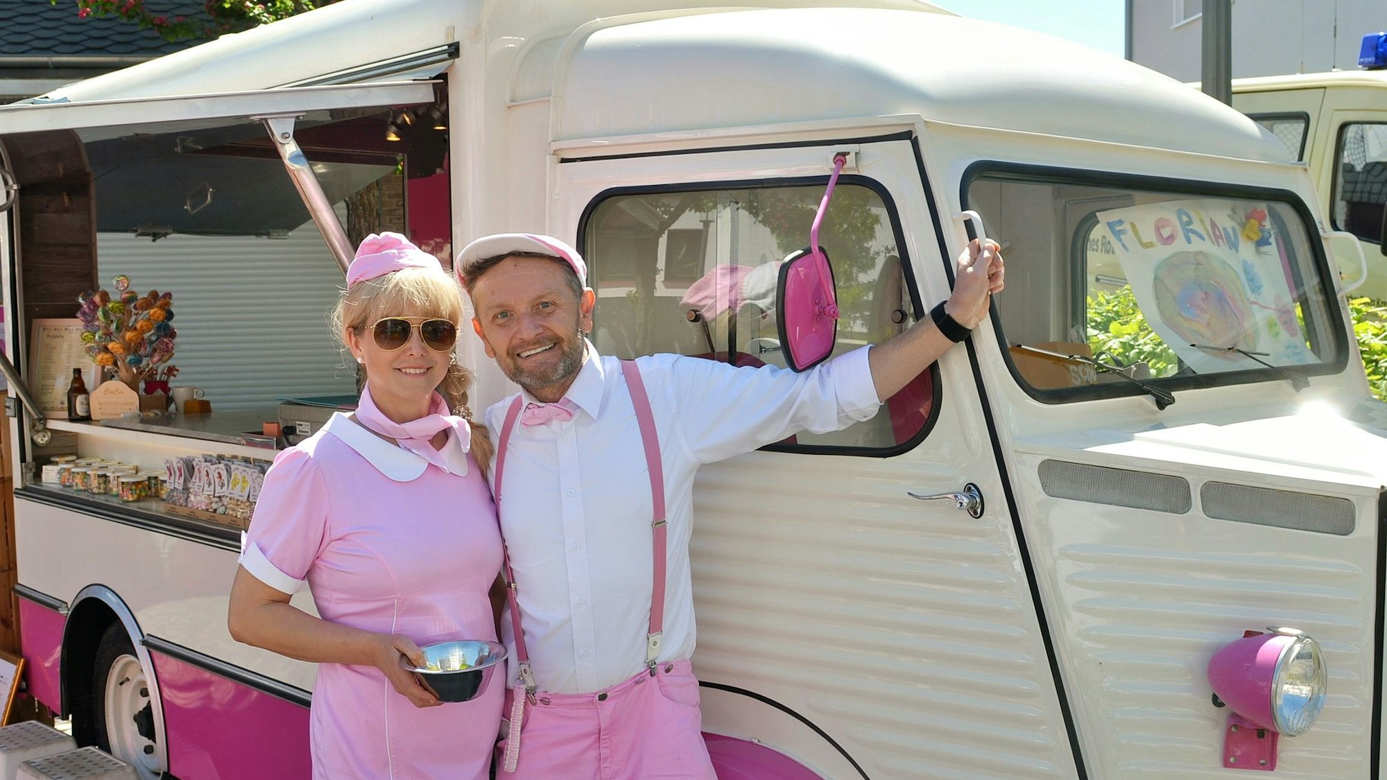 Ein rosa-weiß gekleidetes Paar steht vor einem Oldimer-Verkaufswagen, in dem selbstgemachte Bonbons verkauft werden.