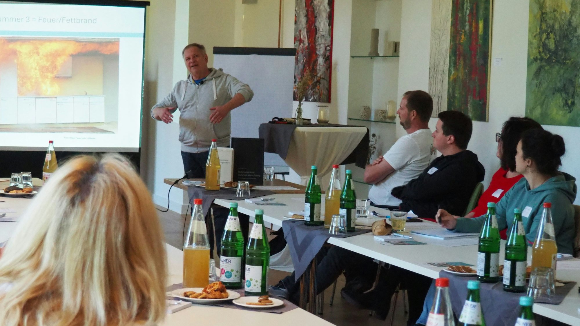 Der Referent spricht vor den Zuhörern. Hinter ihm wird das Bild eines brennenden Gebäudes auf der Leinwand gezeigt.