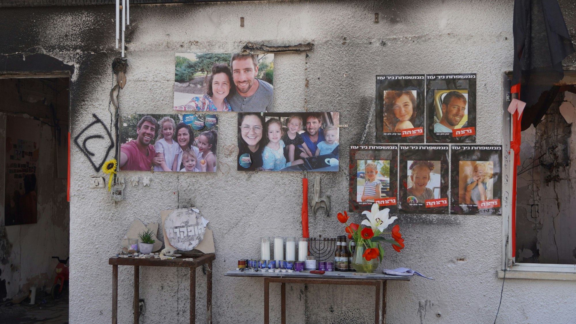 Bilder von Familien und Porträts einzelner Kinder und junger Männer hängen an einer rußgeschwärzten Häuserwand, davor sind Tische mit Kerzen, Blumen und jüdischen Kerzenleuchtern.