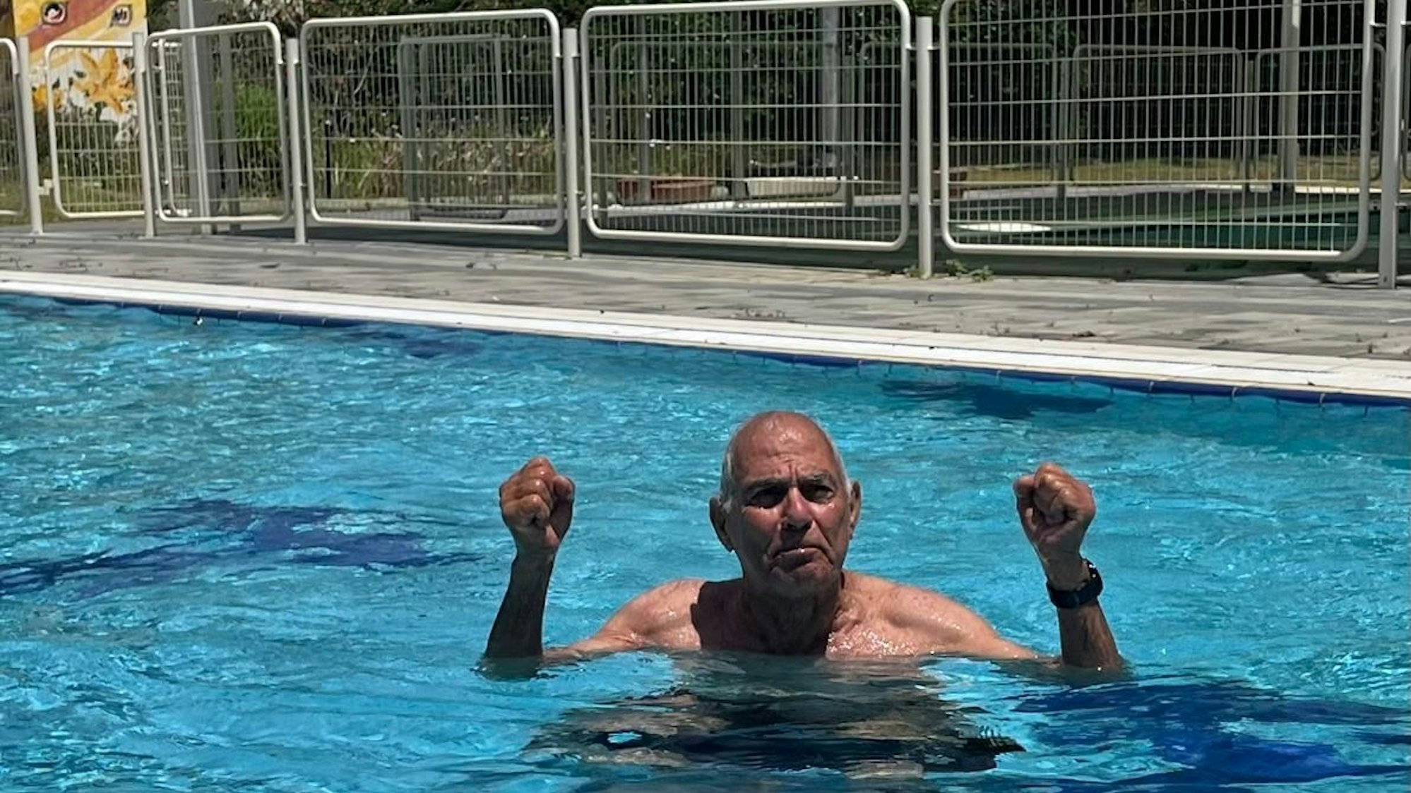 Ein älterer Mann steht in einem Swimming Pool, er reckt die Arme mit geballten Fäusten seitwärts nach oben.
