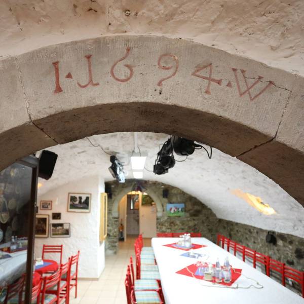 Blick in einen Gewölbekeller, in dem rote Stühle und mit weißen Tischdecken versehene Tische stehen.