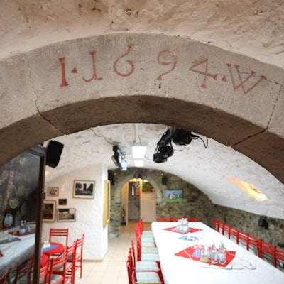 Blick in einen Gewölbekeller, in dem rote Stühle und mit weißen Tischdecken versehene Tische stehen.