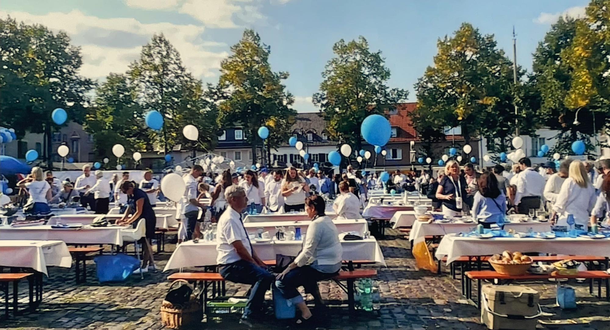 Auf einem Marktplatz stehen zahlreiche Biertische, es gibt Luftbalons in Blau und Weiß, Menschen sitzen an den Tischen.