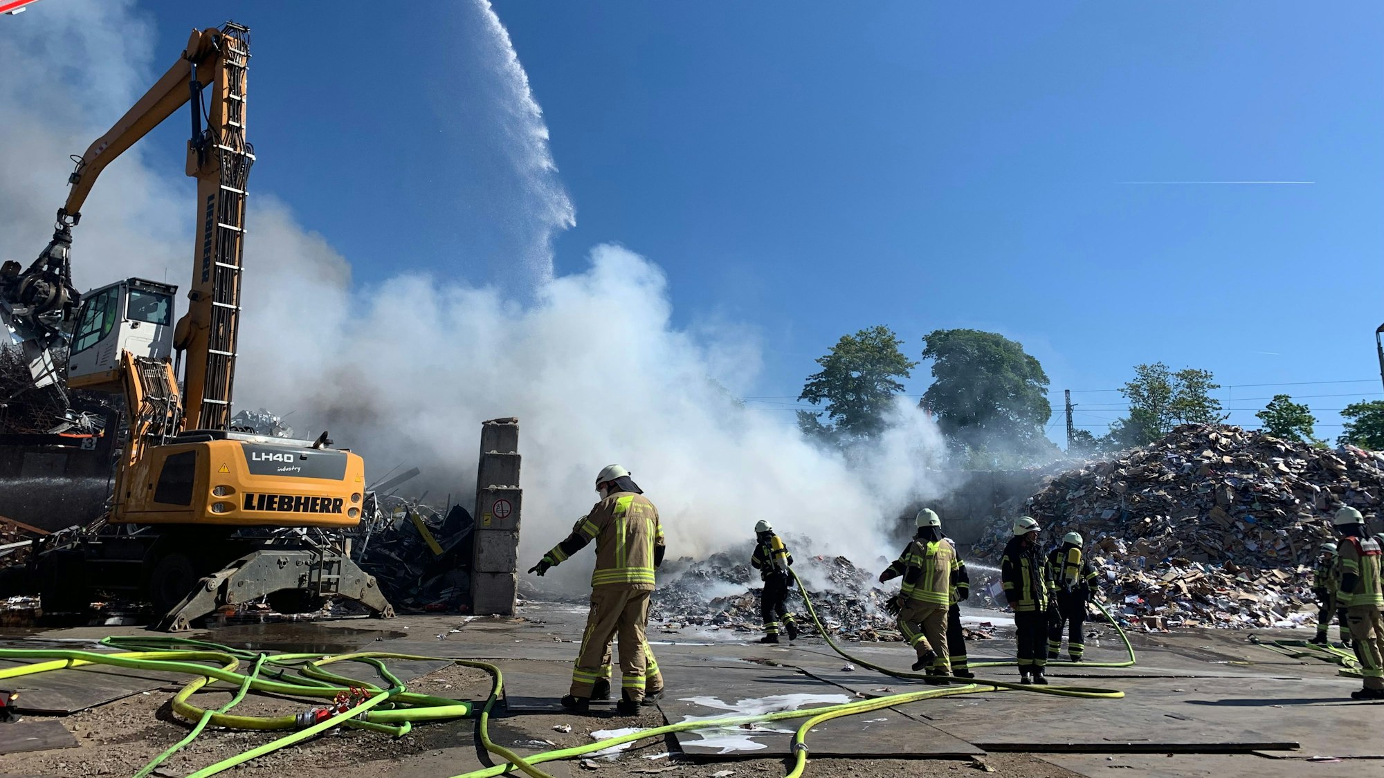 Einsatzkräfte der Feuerwehr und eine Drehleiter vor einer Rauchwolke über einem Müllberg. Links ein Baustellenfahrzeug.