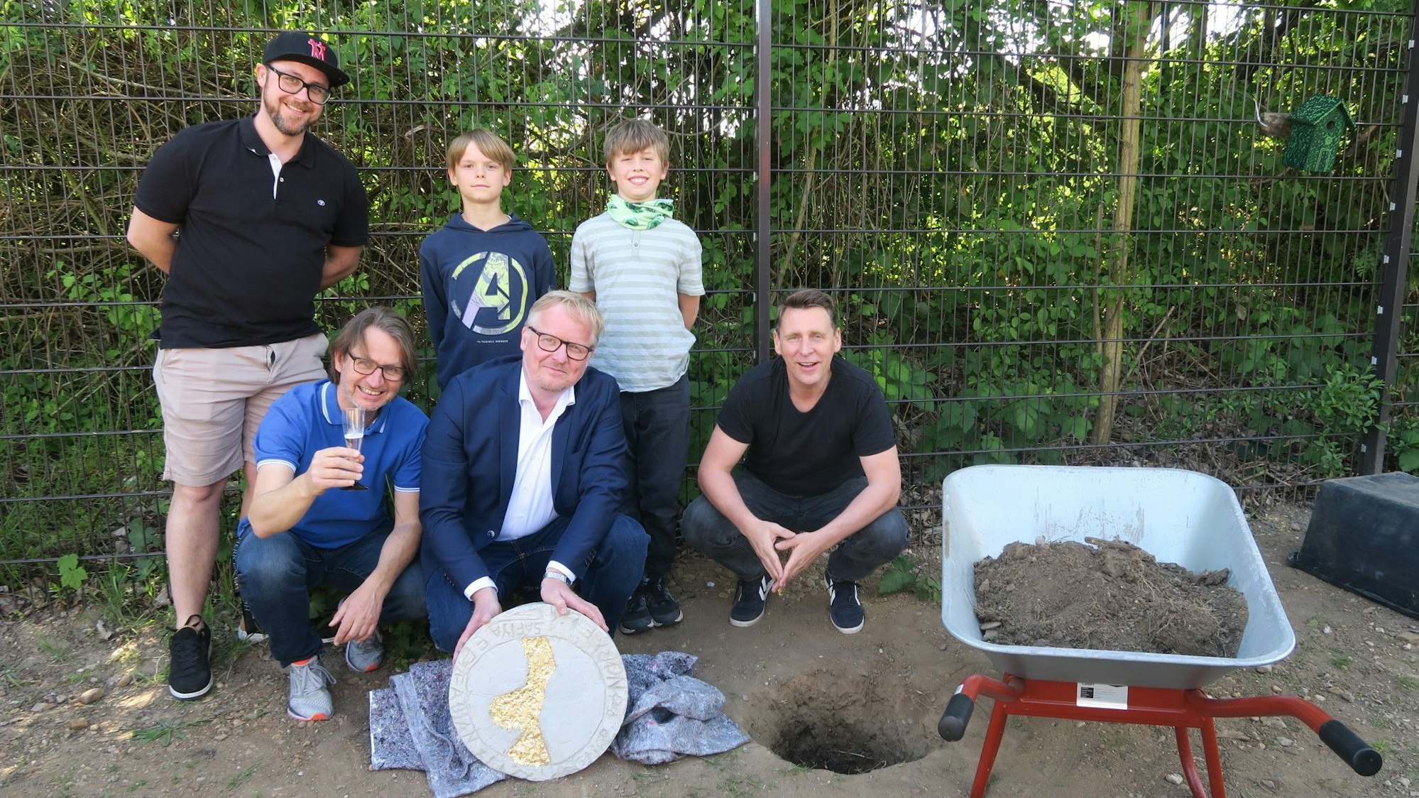 Vier Männer und zwei Jungs stehen zusammen, einer hält die Zeitkapsel, die gleich in ein Loch in der Erde versenkt wird, daneben steht eine Schubkarre voll Erde.