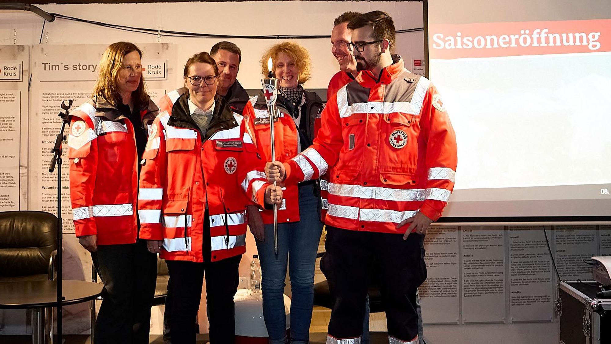 Eine Gruppe Rotkreuzler steht auf der Bühne, zwei halten hält das Rotkreuzlicht in den Händen, ein brennendes Licht an einem langen Metallstab.