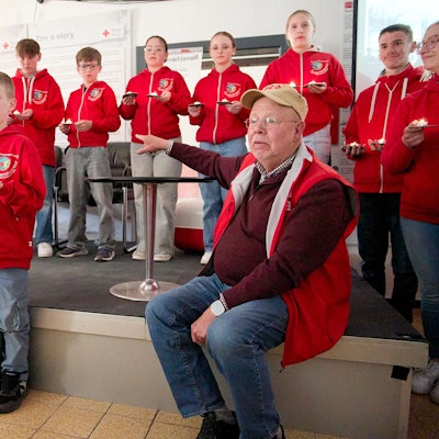 Auf der Bühne stehen Kinder und Jugendliche, davor sitzt auf einem Stuhl Rolf Zimmermann. Alle tragen rote DRK-Kleidung.