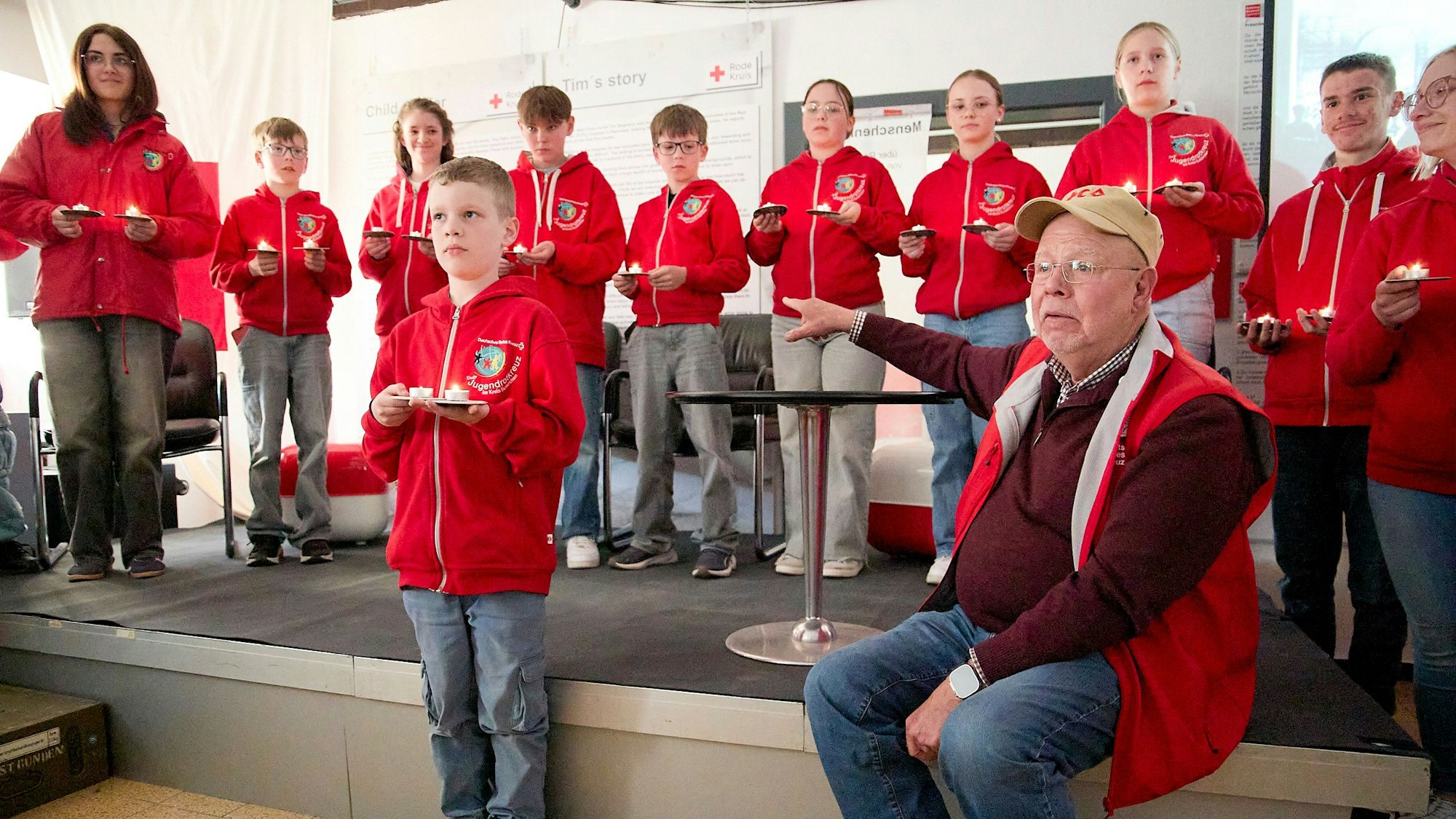 Auf der Bühne stehen Kinder und Jugendliche, davor sitzt auf einem Stuhl Rolf Zimmermann. Alle tragen rote DRK-Kleidung.