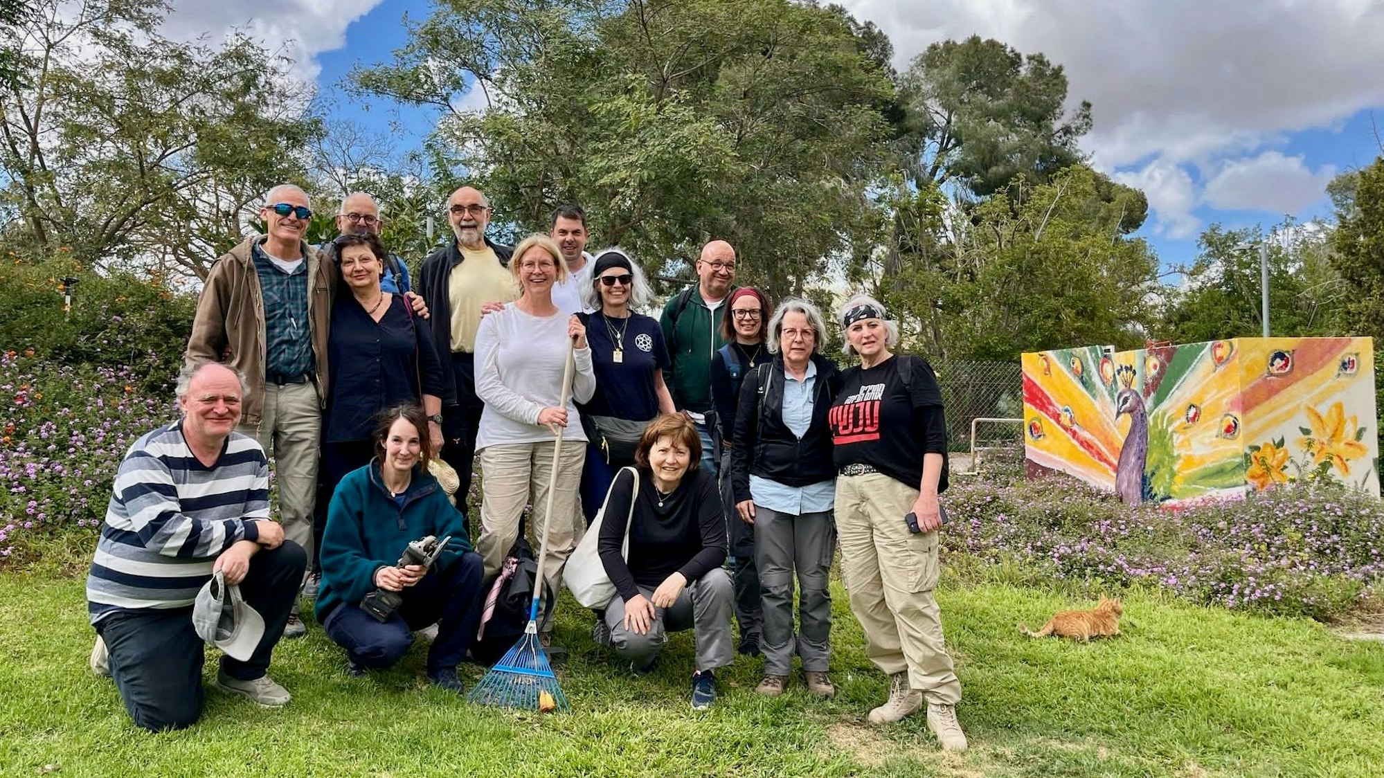 Eine Gruppe von Menschen steht auf einer Wiese, im Hintergrund ist eine mit einem Pfau bemalte Wand zu sehen.