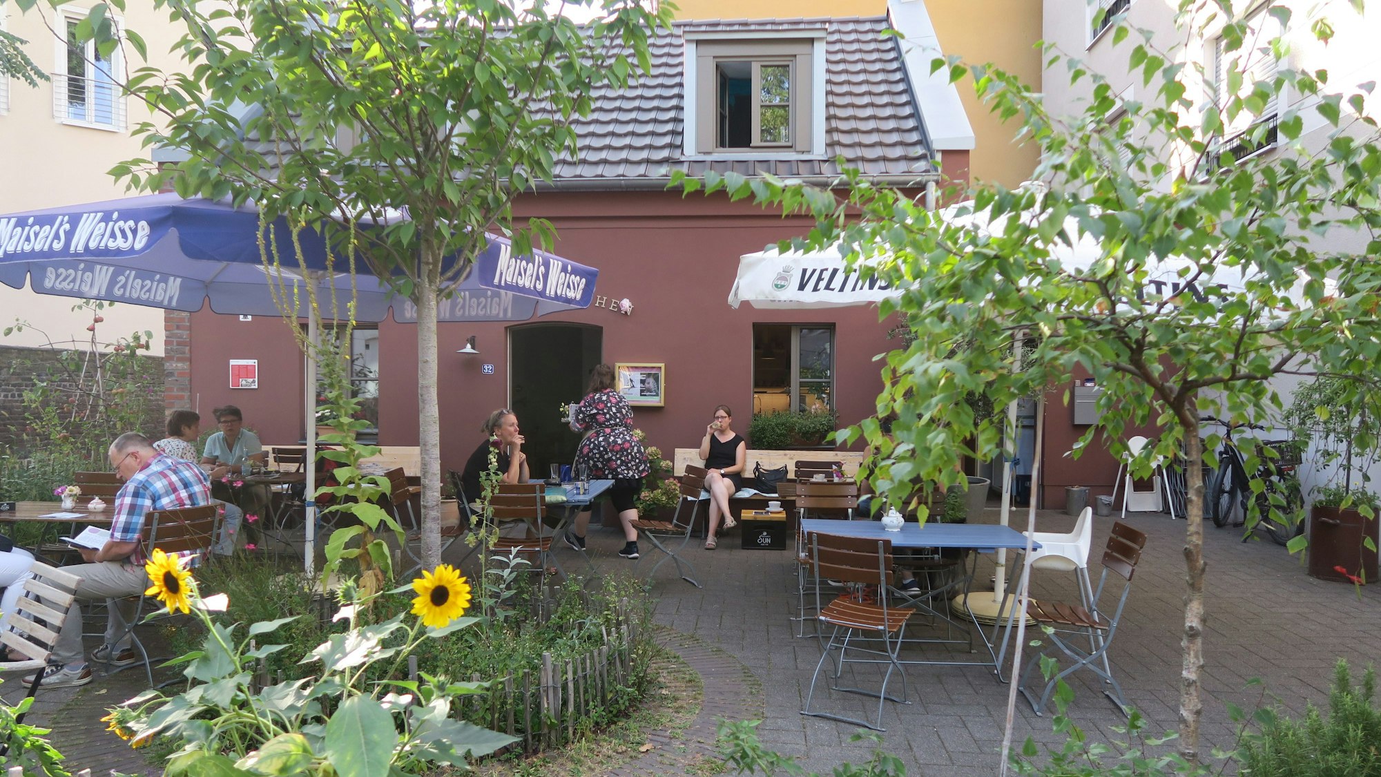 Café-Tische stehen in einem von Pflanzen und einem Holzzaun umrahmten Platz vor einem roten Häuschen.