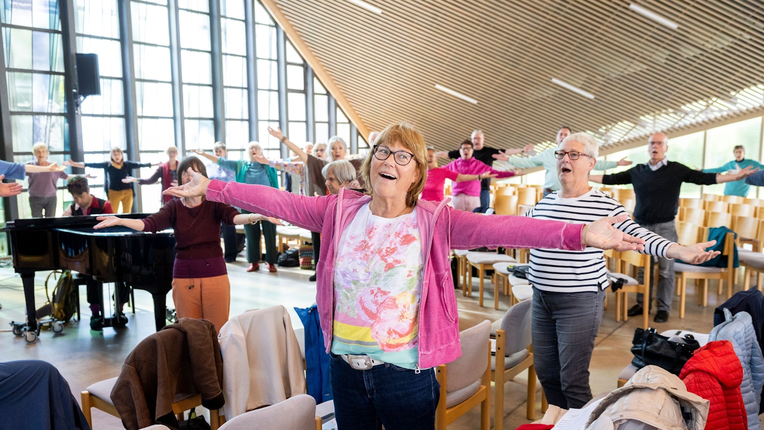 Eine Seniorin hat die Arme ausgebreitet und ist singend zu sehen. Hinter ihr der Chor und ein Pianist in action.