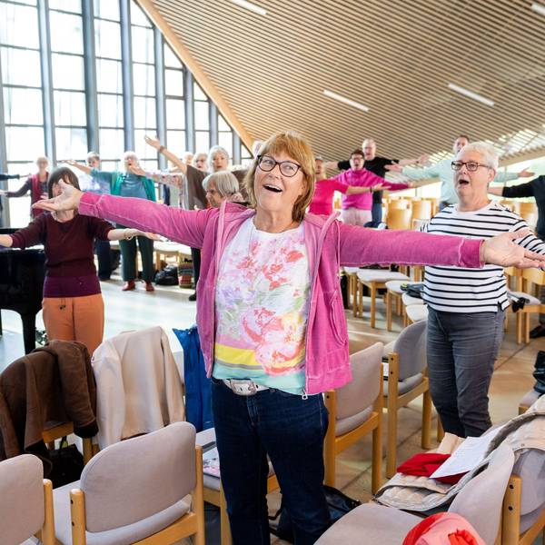 Eine Seniorin hat die Arme ausgebreitet und ist singend zu sehen. Hinter ihr der Chor und ein Pianist in action.