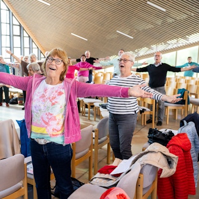 Eine Seniorin hat die Arme ausgebreitet und ist singend zu sehen. Hinter ihr der Chor und ein Pianist in action.