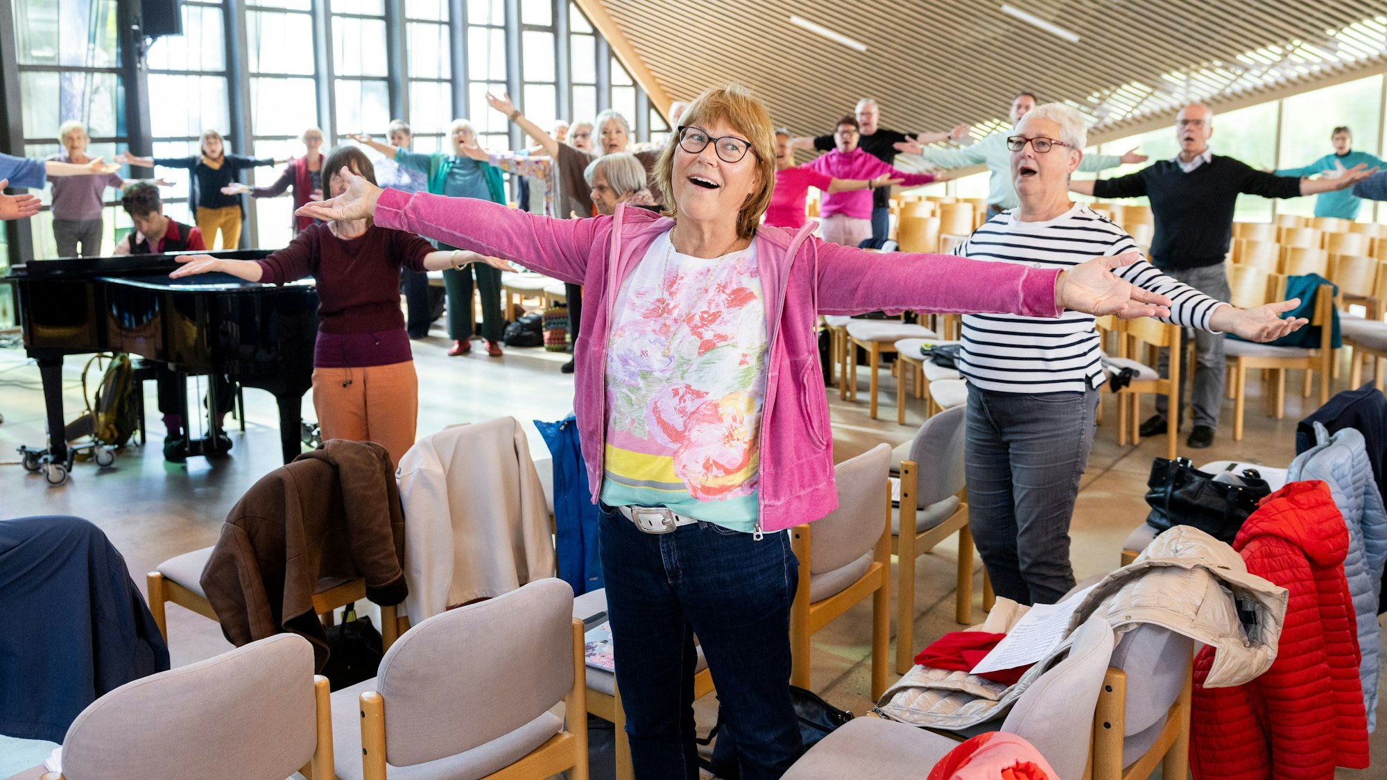 Eine Seniorin hat die Arme ausgebreitet und ist singend zu sehen. Hinter ihr der Chor und ein Pianist in action.