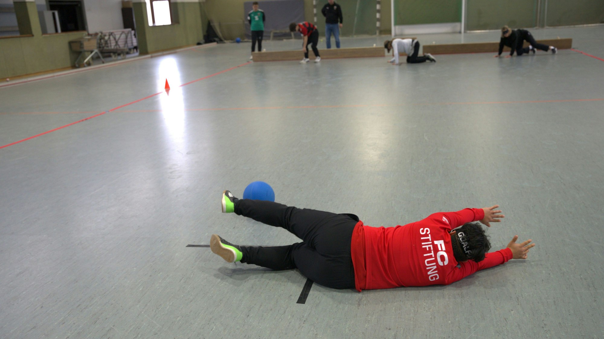 Auf dem Bild ist ein Sportler zu sehen, der auf dem Boden einer Halle liegt und einen Ball abwehrt.