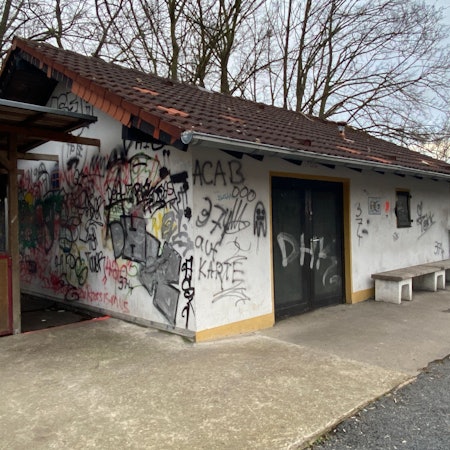 Man sieht ein kleines Haus, das mit vielen Schmierereien überzogen ist und an der linken Seite einen hölzernen Anbau hat.