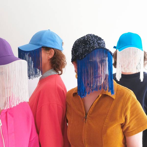 Vier Frauen tragen bunte Oberteile und Baseballkappen, an deren Schirme schleierartige Vorhänge genäht sind
