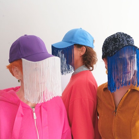 Vier Frauen tragen bunte Oberteile und Baseballkappen, an deren Schirme schleierartige Vorhänge genäht sind