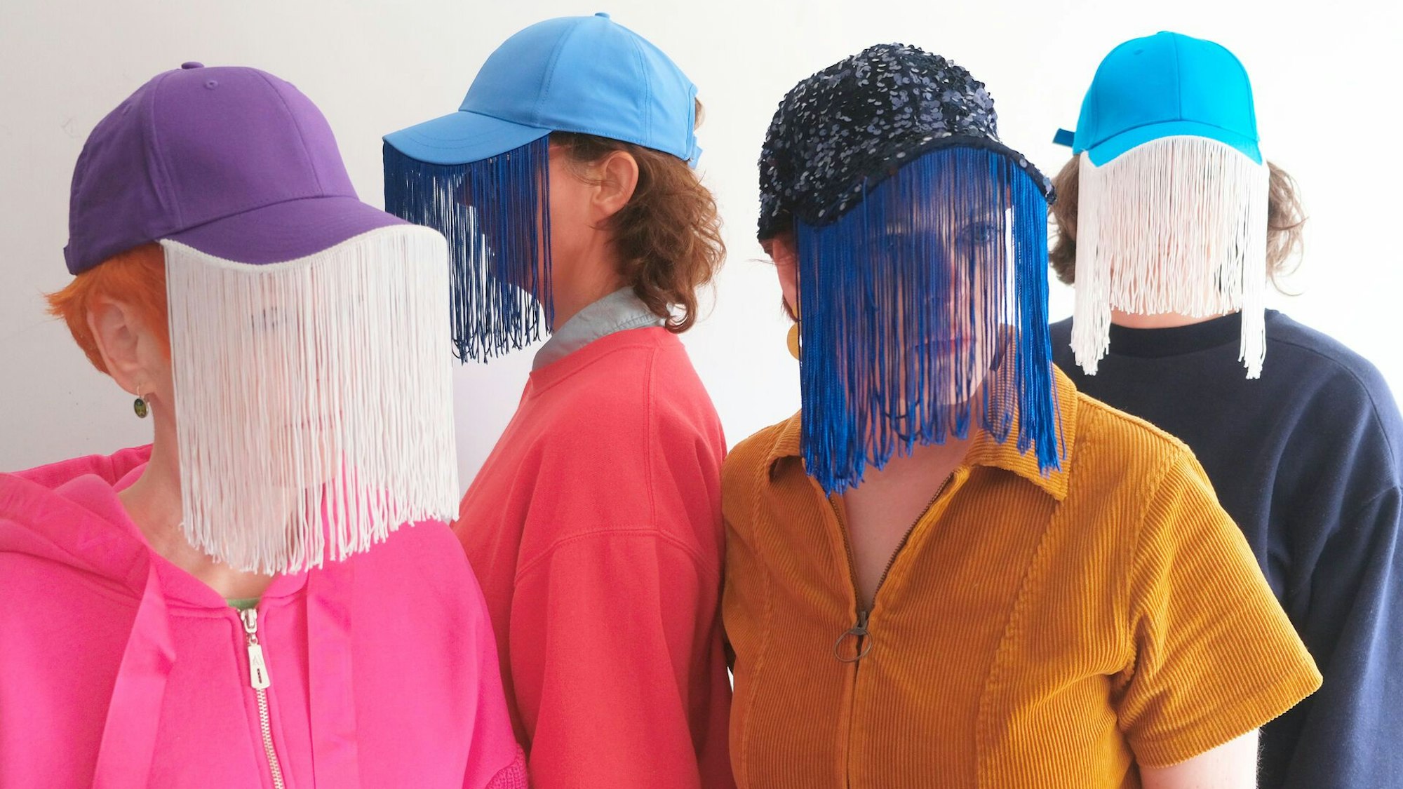 Vier Frauen tragen bunte Oberteile und Baseballkappen, an deren Schirme schleierartige Vorhänge genäht sind
