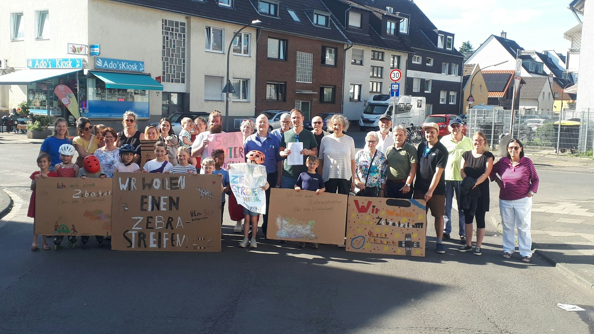 Eltern und Kinder, sowie Vertreter von Niehler Bürgerverein und der Bezirksvertretung Nippes, stehen auf einer Straße und fordern mit Plakaten einen Zebrastreifen an der Merkenicher Straße, um den Schulweg zur GGS Halfengasse sicherer zu machen.
