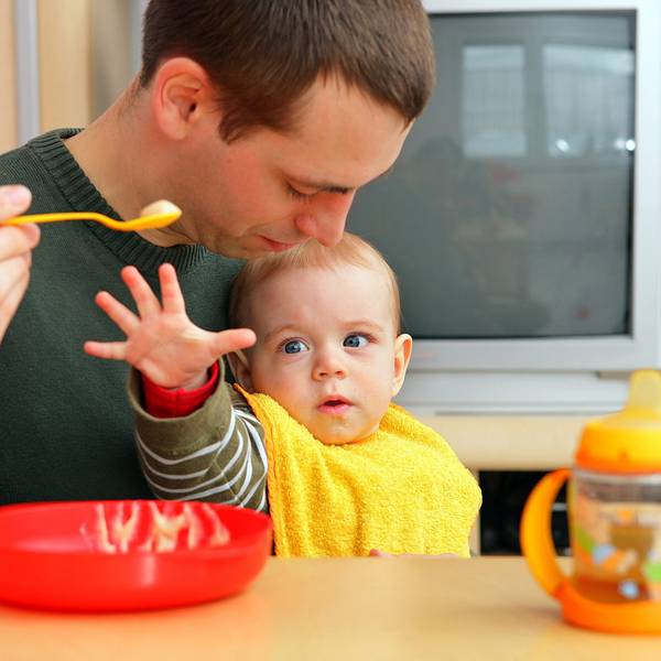Ein Vater füttert sein Baby auf dem Schoß. Das greift nach dem Breilöffel.