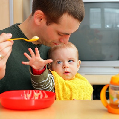 Ein Vater füttert sein Baby auf dem Schoß. Das greift nach dem Breilöffel.