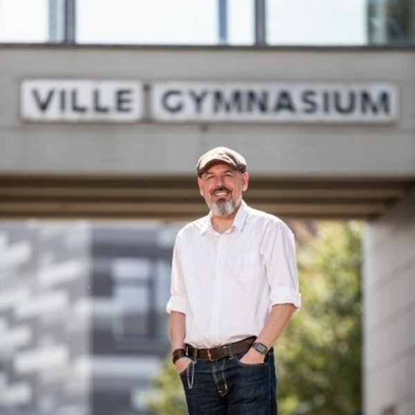 Auf dem Foto ist ein Mann mit Vollbart in weißem Hemd und einer Schiebermütze zu sehen.