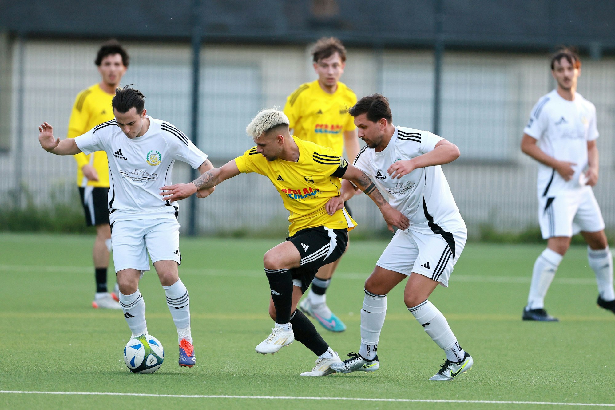 Das Foto zeigt einen Bachemer Spieler im gelben Trikot, der von zwei Glescher Gegenspielern (in Weiß) nicht an den Ball gelassen wird.