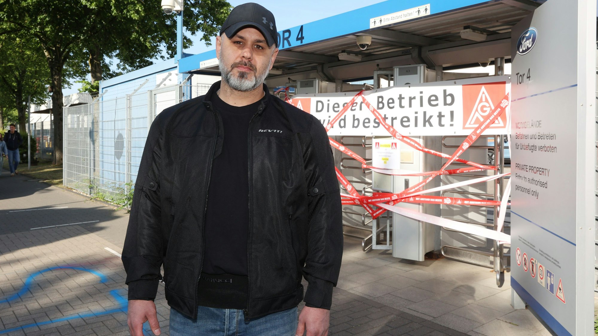 Joachim Reitmeier (43 Jahre) trägt eine schwarze Jacke und ein Basecap. Er steht vor einem gesperrten Zugang zum Ford-Werk. Foto: Martina Goyert