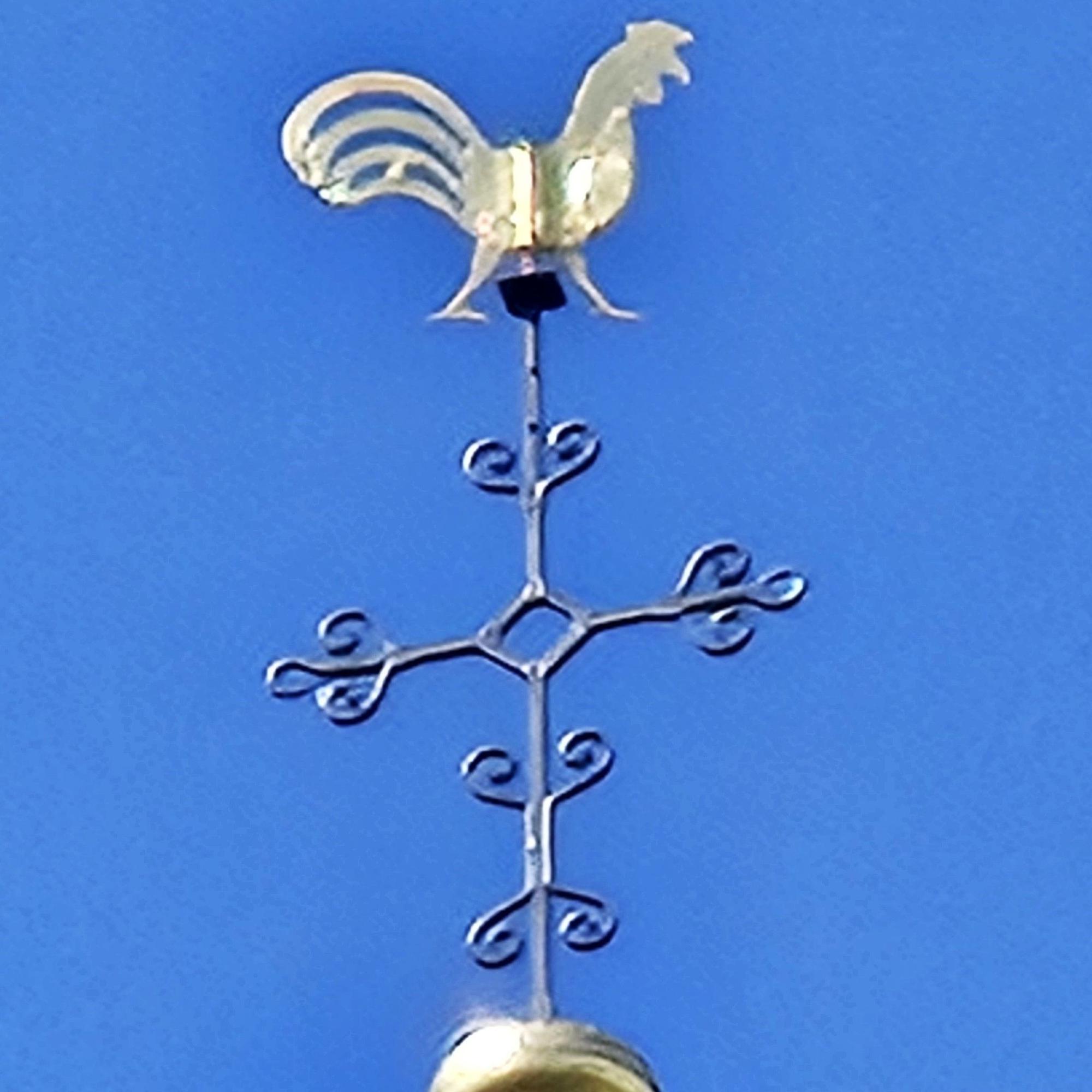Ein goldener Hahn thront über einem stilisierten Kreuz.