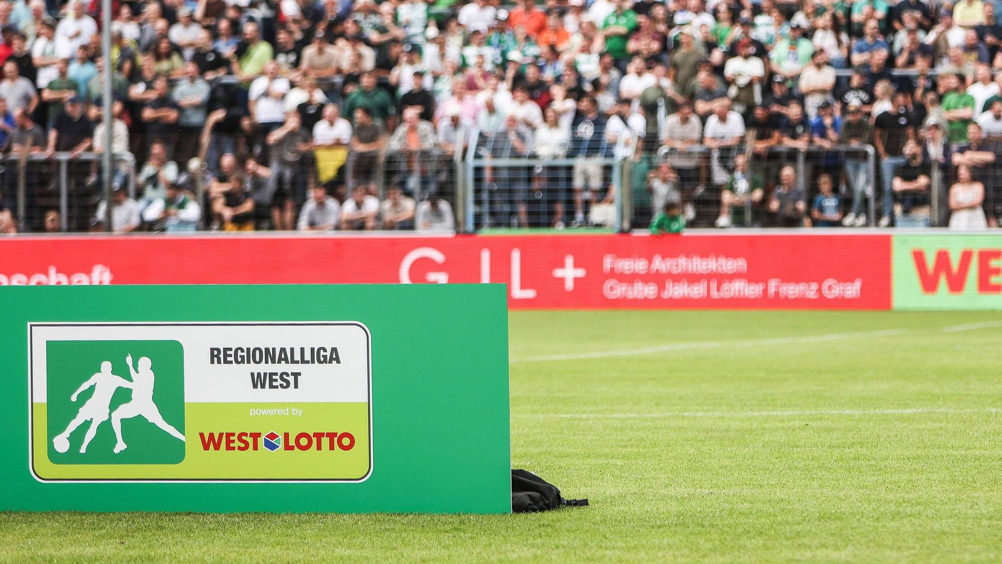 Vor einer Fankurve in einem Fußballstadion steht ein Schild mit der Aufschrift „Regionalliga West“.