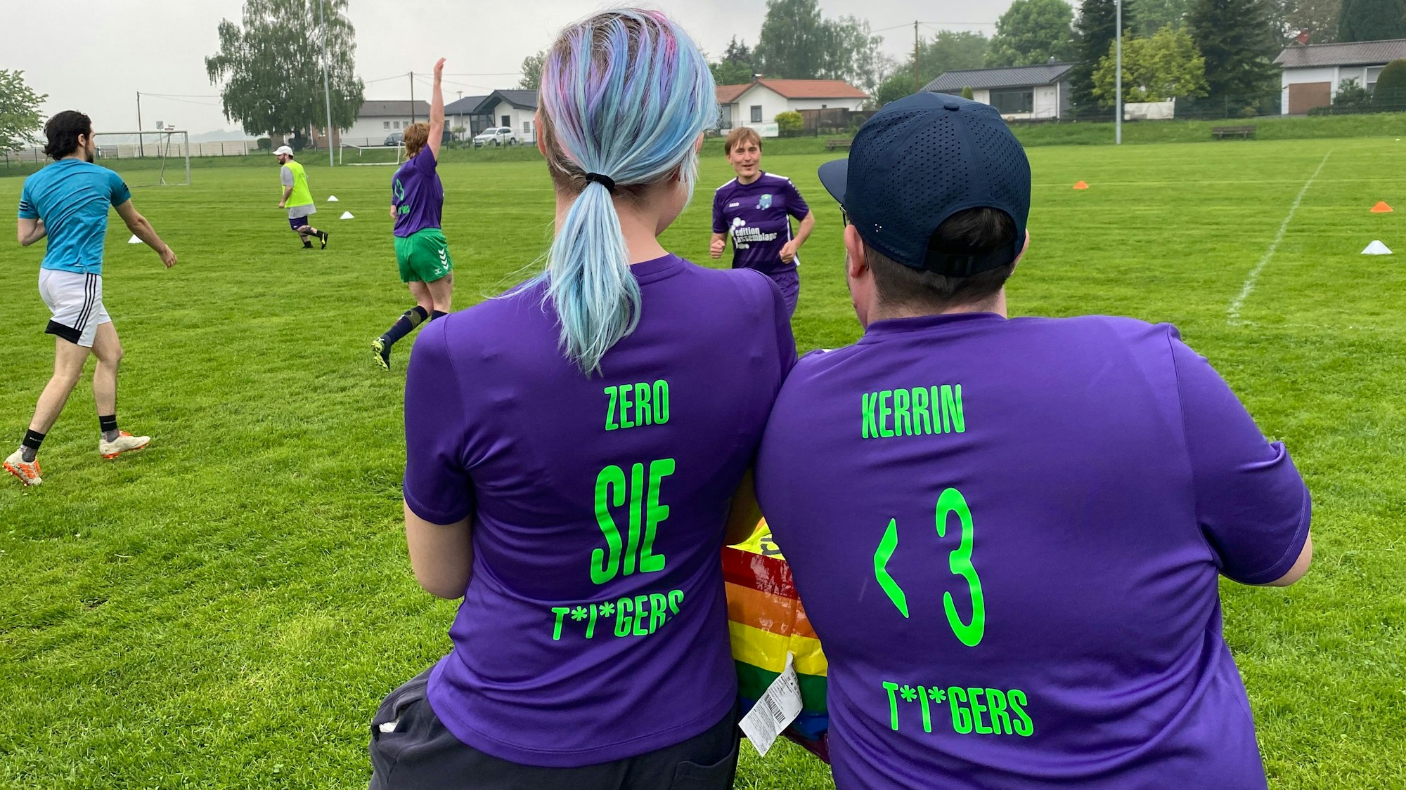 Zwei Personen in lilafarbenen Tigers-Trikots stehen mit dem Rücken zur Kamera. Im Hintergrund sieht man andere aus der Gruppe auf dem Fußballplatz kicken.