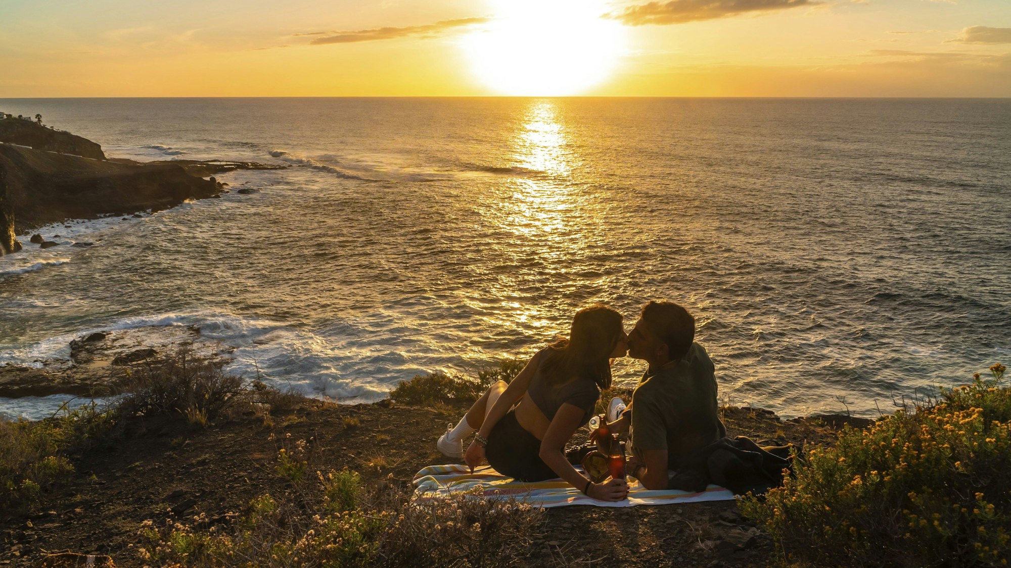 Romantisches Paar, das sich küsst, während es Getränke und den Sonnenuntergang über dem Meer in Agaete, Gran Canaria genießt.