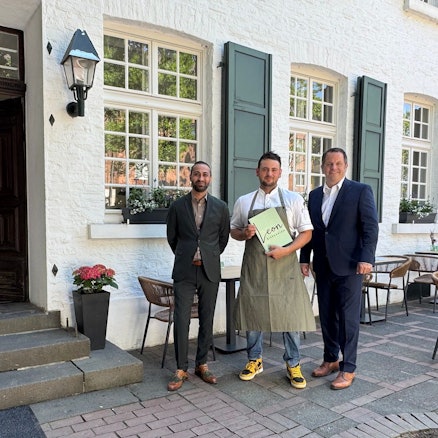 Drei Männer stehen vor einem Restaurant, der in der Mitte hält eine Speisekarte mit dem Schriftzug „Leon“ in den Händen.