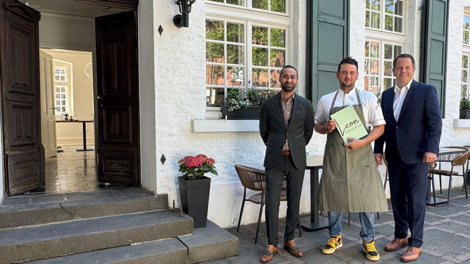 Drei Männer stehen vor einem Restaurant, der in der Mitte hält eine Speisekarte mit dem Schriftzug „Leon“ in den Händen.
