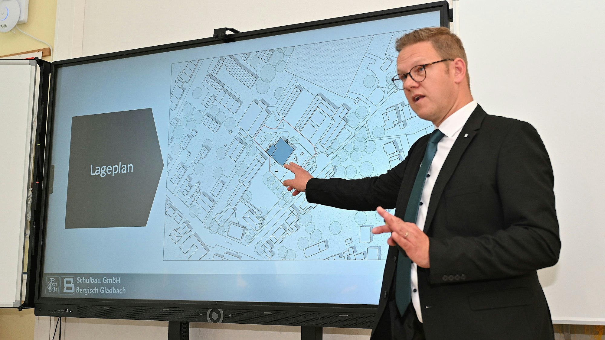 Rolko steht vor einer Leinwand, auf der der Lageplan für den Neubau abgebildet ist.