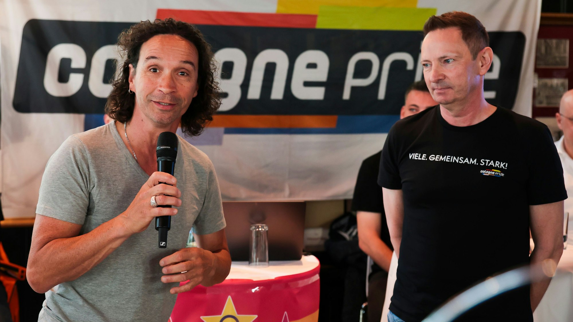 Stefan Brings (links) hält ein Mikrofon in der Hand und schaut in die Kamera. Rechts neben ihm steht ColognePride-Geschäftsführer Uwe Weiler.