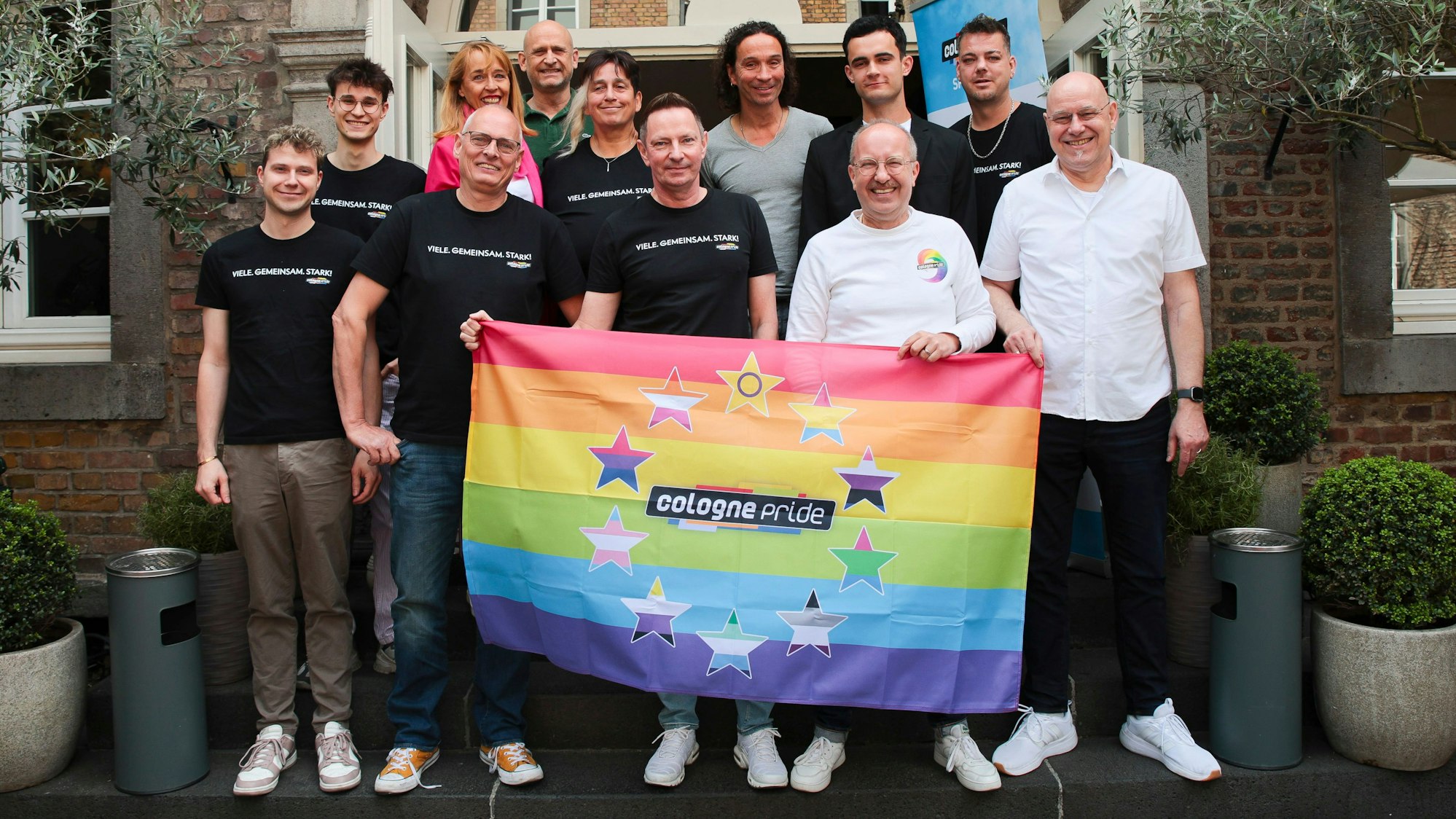 Zu sehen ist das Team der Cologne Pride auf einem Gruppenfoto. Drei Personen halten eine Regenbogenfahne.